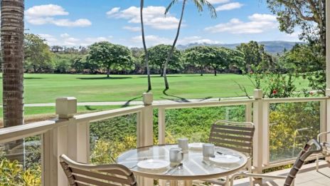 A sunny balcony with a round table and chairs overlooking a lush green field and palm trees, blue sky with fluffy clouds, distant hills.