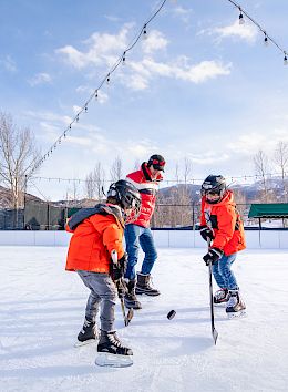 The image shows a man coaching two children in orange jackets as they skate and play hockey on an ice rink against a snowy backdrop.
