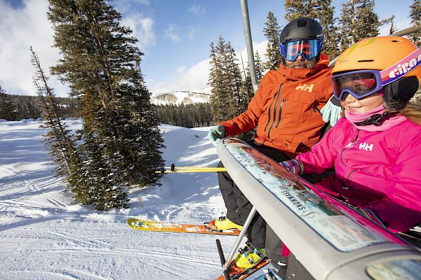A man and a girl ride a ski lift, both wearing helmets and winter gear, surrounded by snowy trees and a mountainous backdrop.