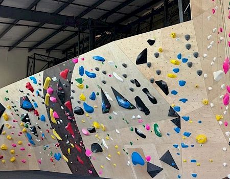 A wide indoor bouldering gym with colorful hold routes on angular white walls and black climbing volumes, lit by ceiling lights, with mats below.