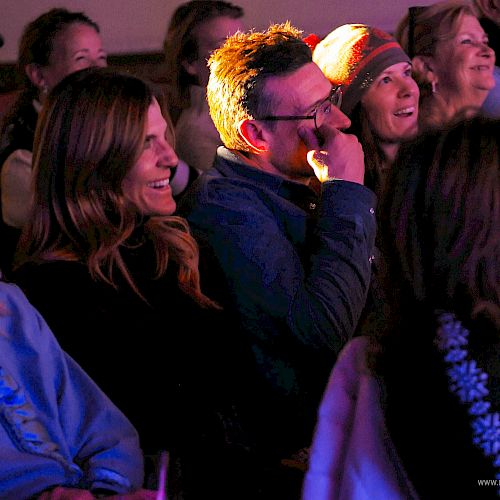 A group of people in a dim, colorful venue watching a performance, smiling and laughing as they sit close together.