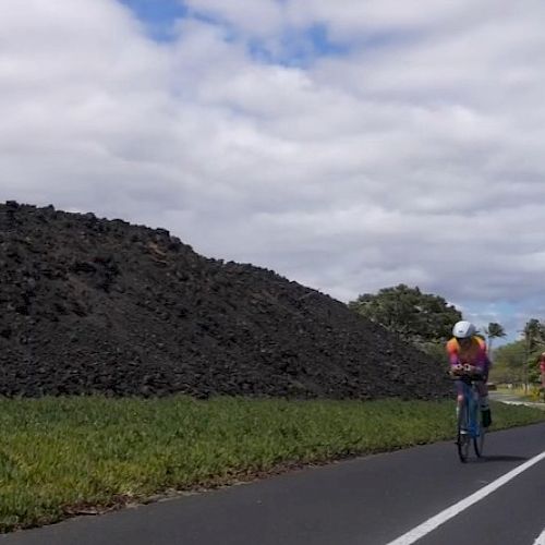 Cyclists ride along a road beside a large dark stone wall and green fields under a cloudy sky.