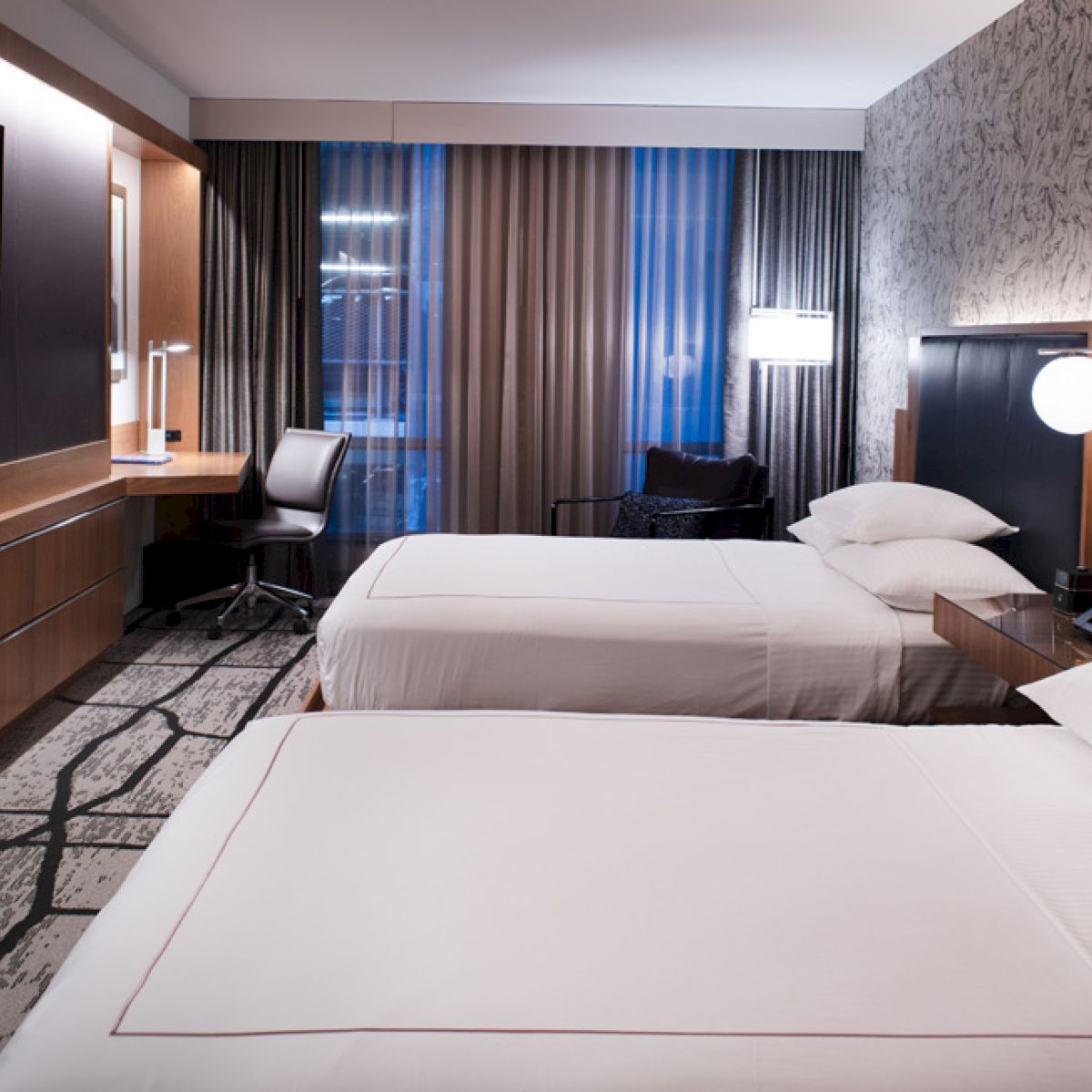 This image shows a modern hotel room with two beds, a flat-screen TV, a desk with a chair, and large windows covered with curtains.