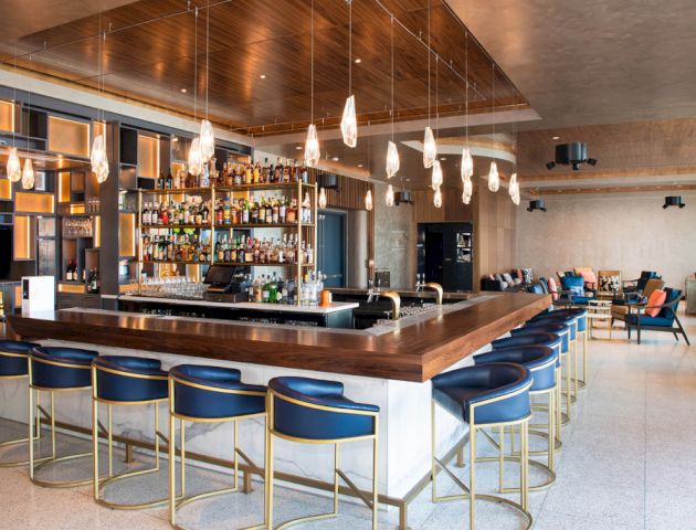 A modern bar with a central counter, blue bar stools, hanging lights, and a well-stocked bar shelf. The background includes seating and decor.