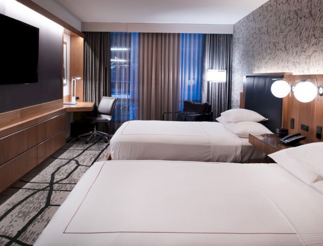 A modern hotel room with two beds, a desk, a flat-screen TV, and contemporary decor including patterned wallpaper and curtains.