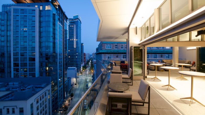 A cityscape view from a modern balcony with seating arrangements, overlooking tall buildings and a street below at dusk.