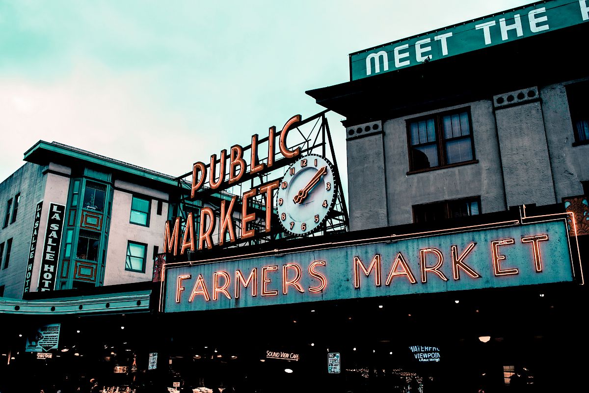 A neon sign reads "Public Market" with a clock and "Farmers Market" below, in a busy market area with surrounding old-style buildings.
