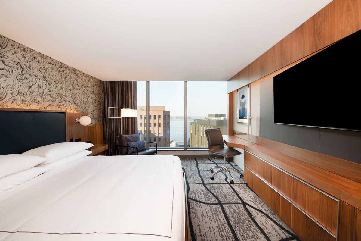 The image shows a modern hotel room with a large bed, a wall-mounted TV, a functional work desk, and a view of the city through large windows.
