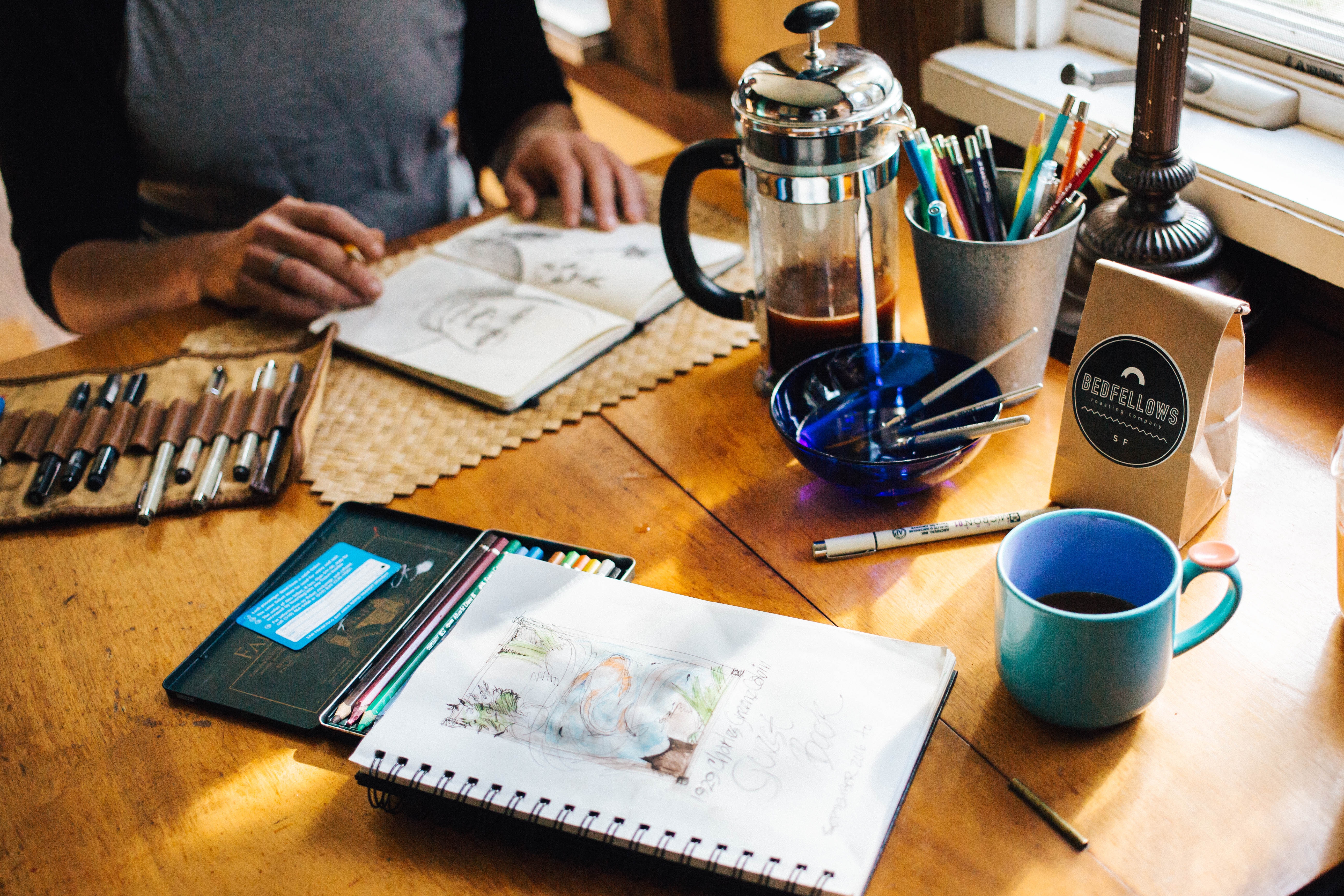 A person sketching at a wooden table with art supplies, French press coffee, a coffee bag, a blue mug, and colored pencils.