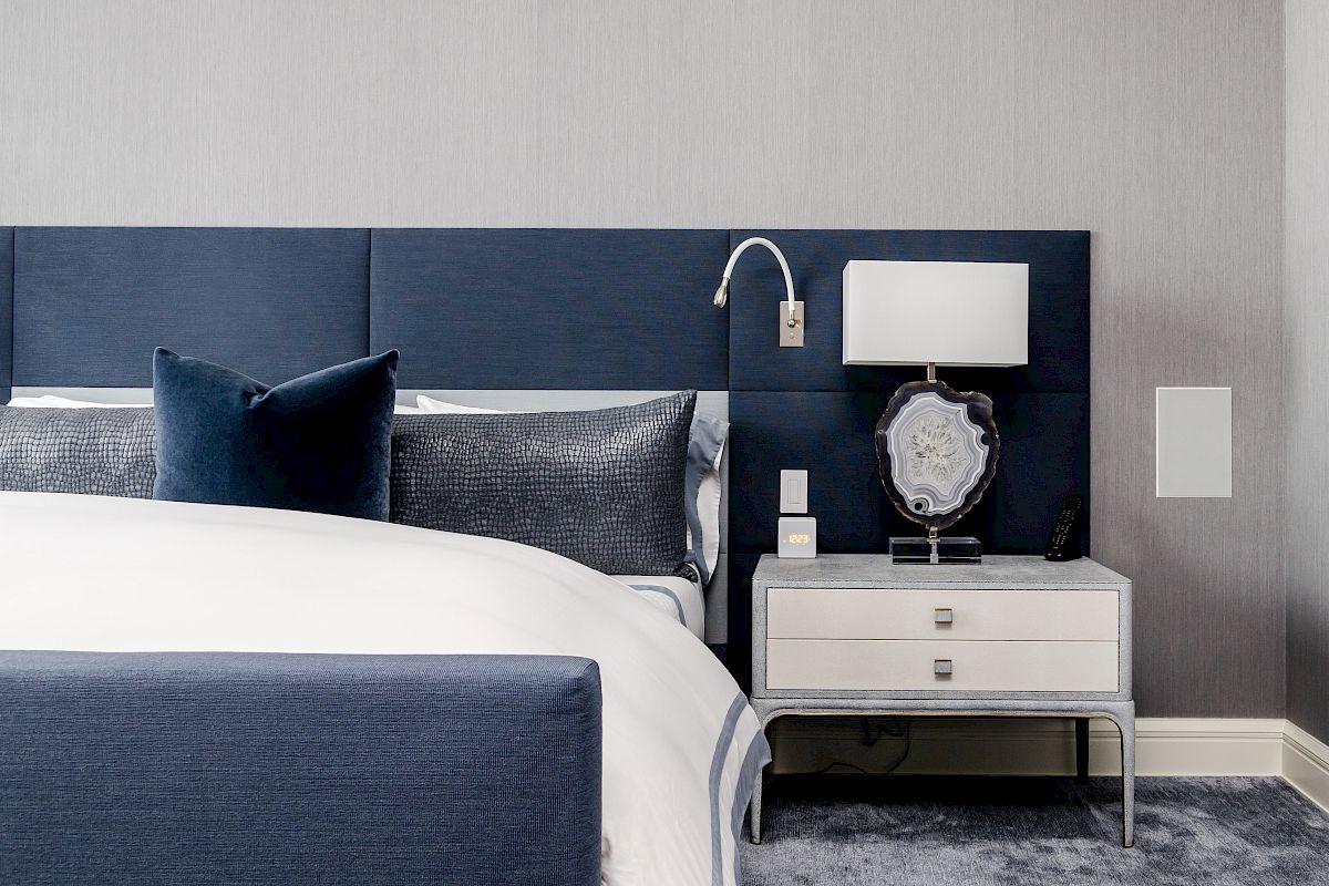 This image shows a modern bedroom with a blue and white color scheme, featuring a bed, pillows, a nightstand with a lamp, and decorative items.